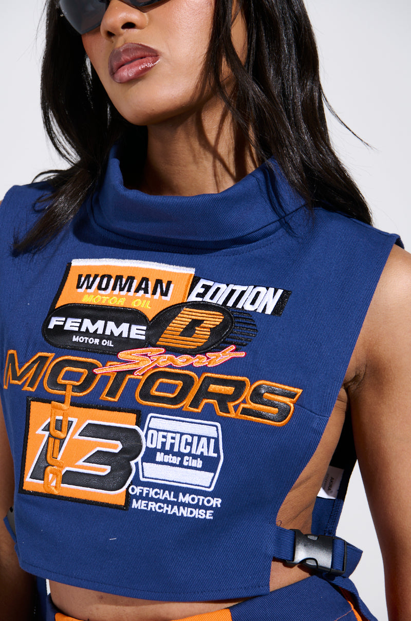 A woman with long hair dons the sleeveless MOTORS TOP, a blue denim piece adorned with vibrant racing-inspired patches such as "Woman Edition," "Motors," and "Official Motor Merchandise." Her sunglasses add a chic touch against the plain white background.