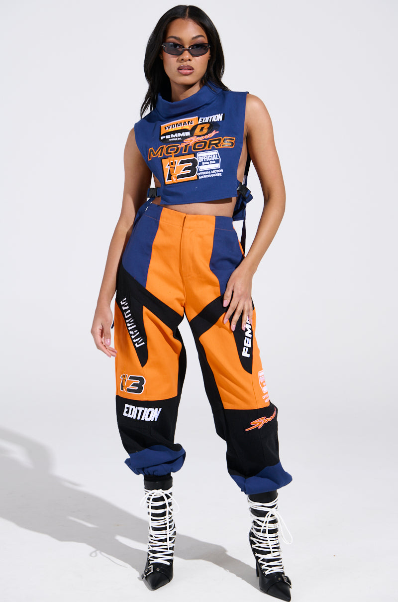 A woman wears racing-inspired clothing with a chic color-block design, featuring orange, blue, and black with bold graphics and patches. She accessorizes with sunglasses and white lace-up boots, exuding confidence akin to the MOTOS ABSTRACT JOGGER.