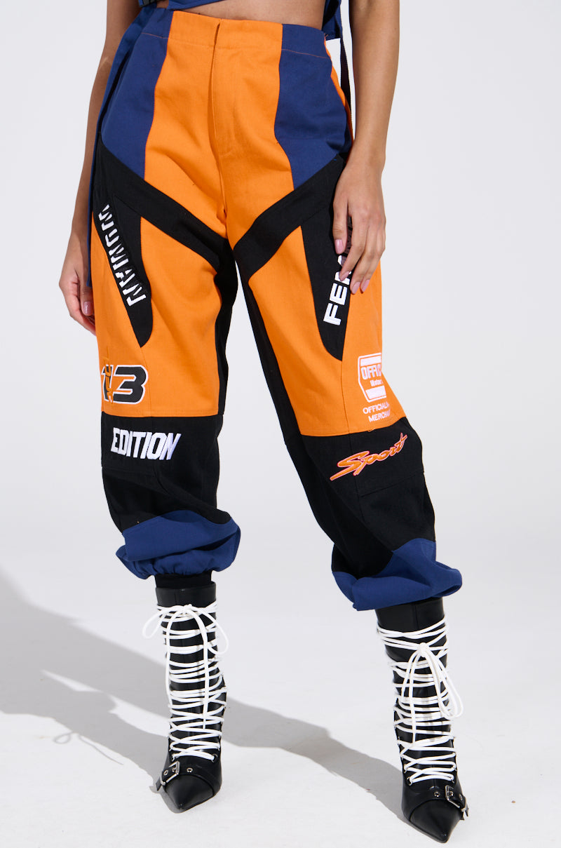 A person wearing MOTOS ABSTRACT JOGGER pants in orange, blue, and black with racing patches and bold angles, paired with white lace-up shoes in an open style against a plain white background.