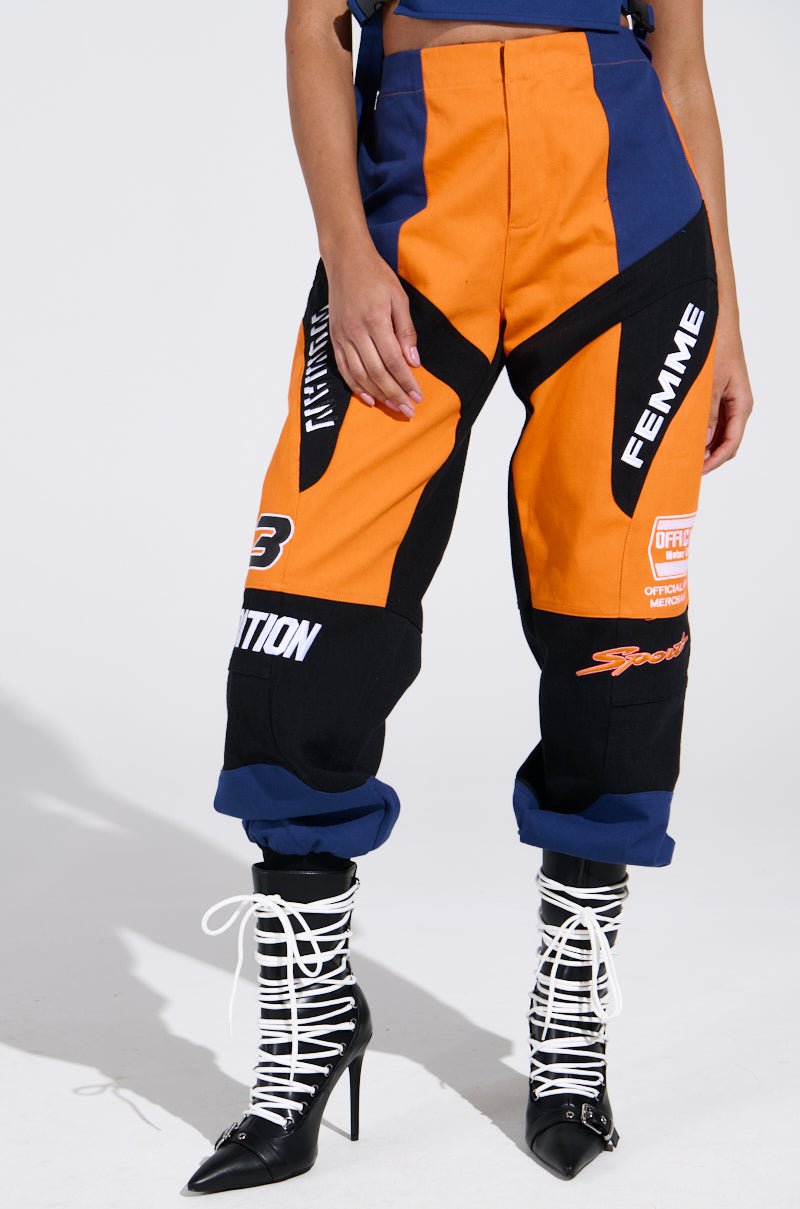 A person wearing the MOTOS ABSTRACT JOGGER pants with a color-block design in orange and black, enhanced by blue accents, poses against a light backdrop. The outfit includes racing-inspired patches and logos. They complement it with black pointed-toe boots featuring white crisscross laces.
