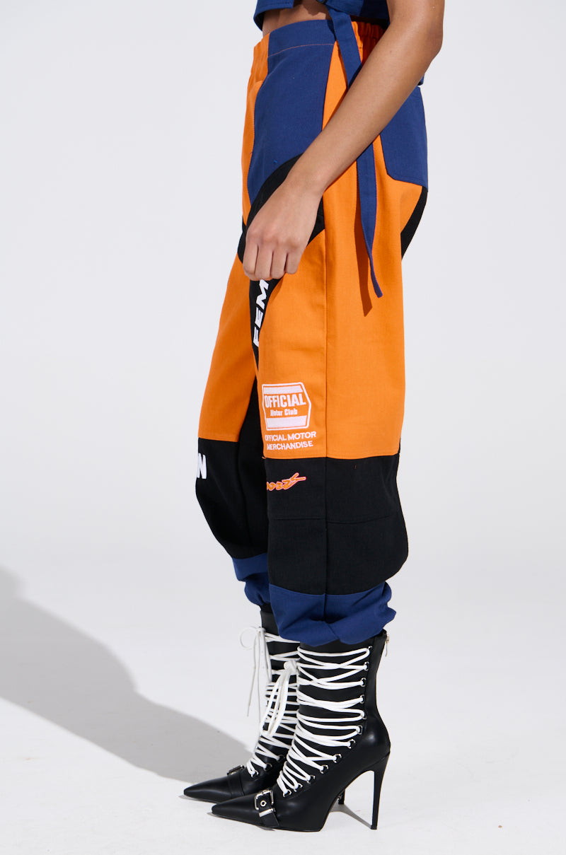 A person wearing the MOTOS ABSTRACT JOGGER in vibrant orange, blue, and black with racing-inspired patches exudes a sporty flair. They also sport white lace-up high-heeled boots with pointed toes against a plain white background.