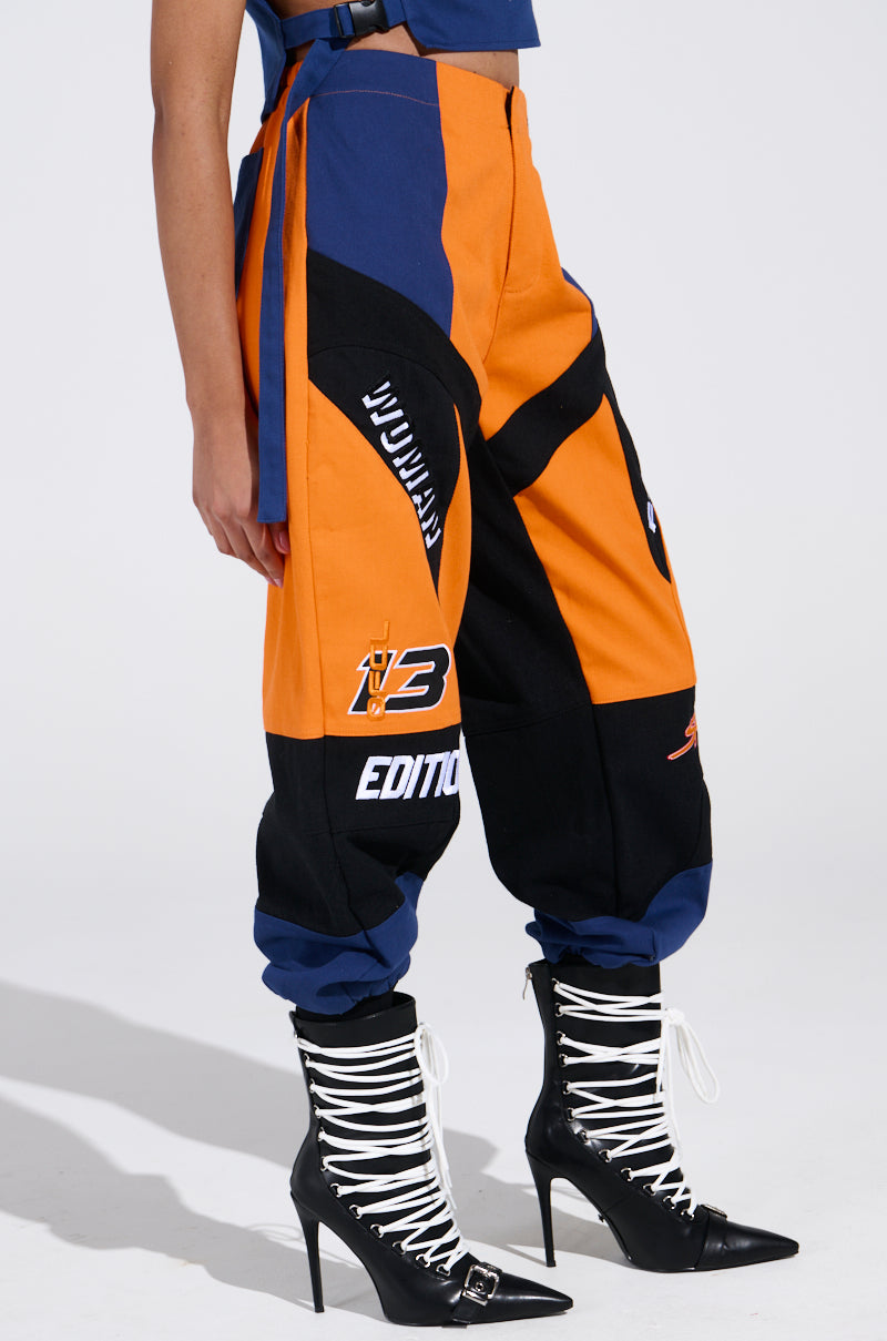 The person wears MOTOS ABSTRACT JOGGER pants in orange and black color-block, featuring white text and logos. They pair them with black high-heeled boots with white laces against a plain white background.