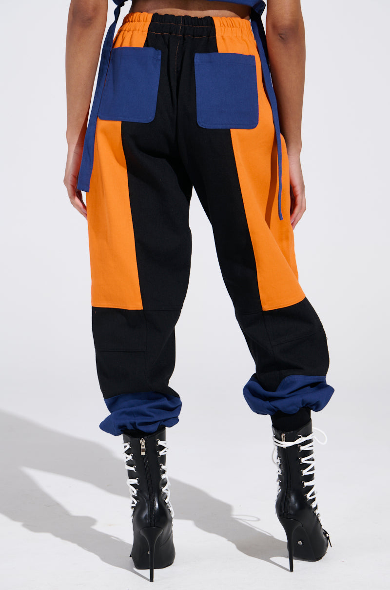 Someone is wearing the MOTOS ABSTRACT JOGGERS in black, orange, and blue with large pockets and a color-block design featuring racing-inspired patches, paired with black lace-up boots against a plain white background.