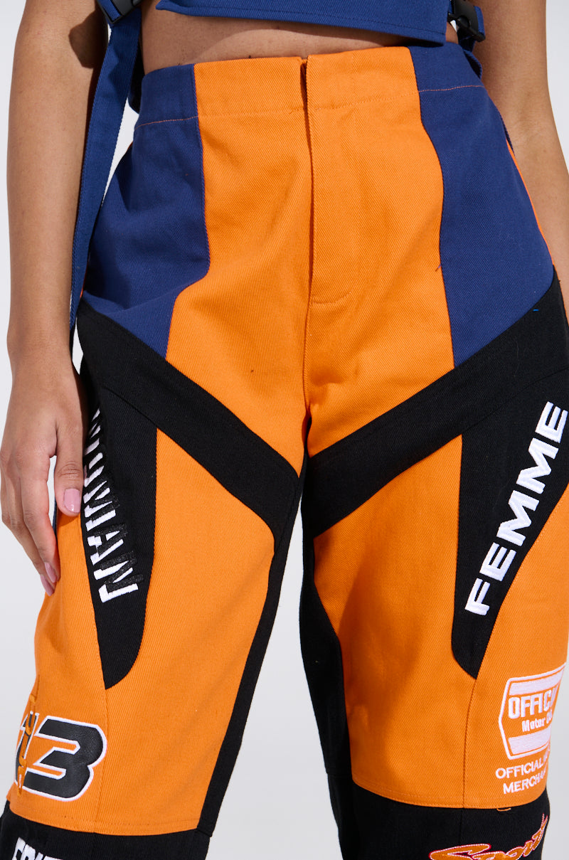 A person wearing the MOTOS ABSTRACT JOGGER, featuring a vibrant racing-inspired color-block design in orange, black, and blue patterns with logos and "FEMME," stands against a plain background.