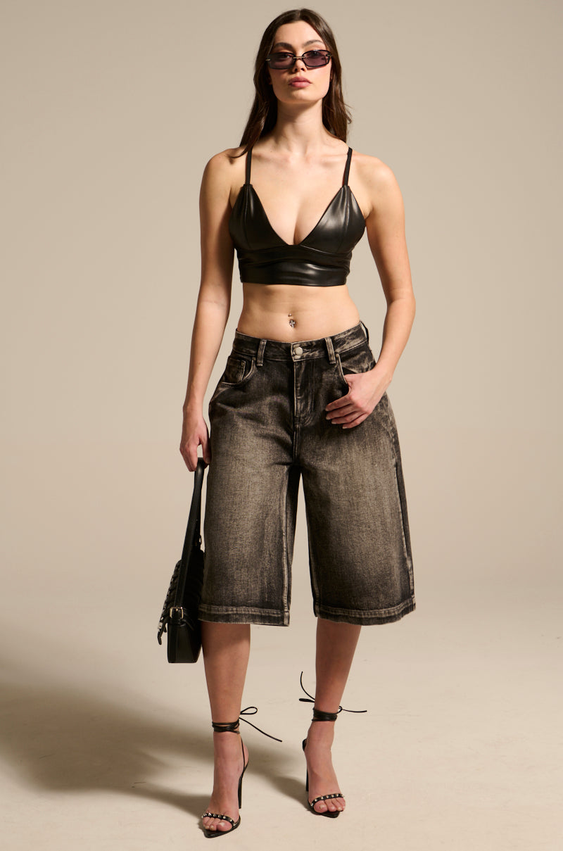 Wearing the PAXTON Sexy Faux Leather Triangle Longline Bralette with knee-length denim shorts, a black handbag, and strappy high heels, a woman radiates confidence while stylishly posing against a neutral background.