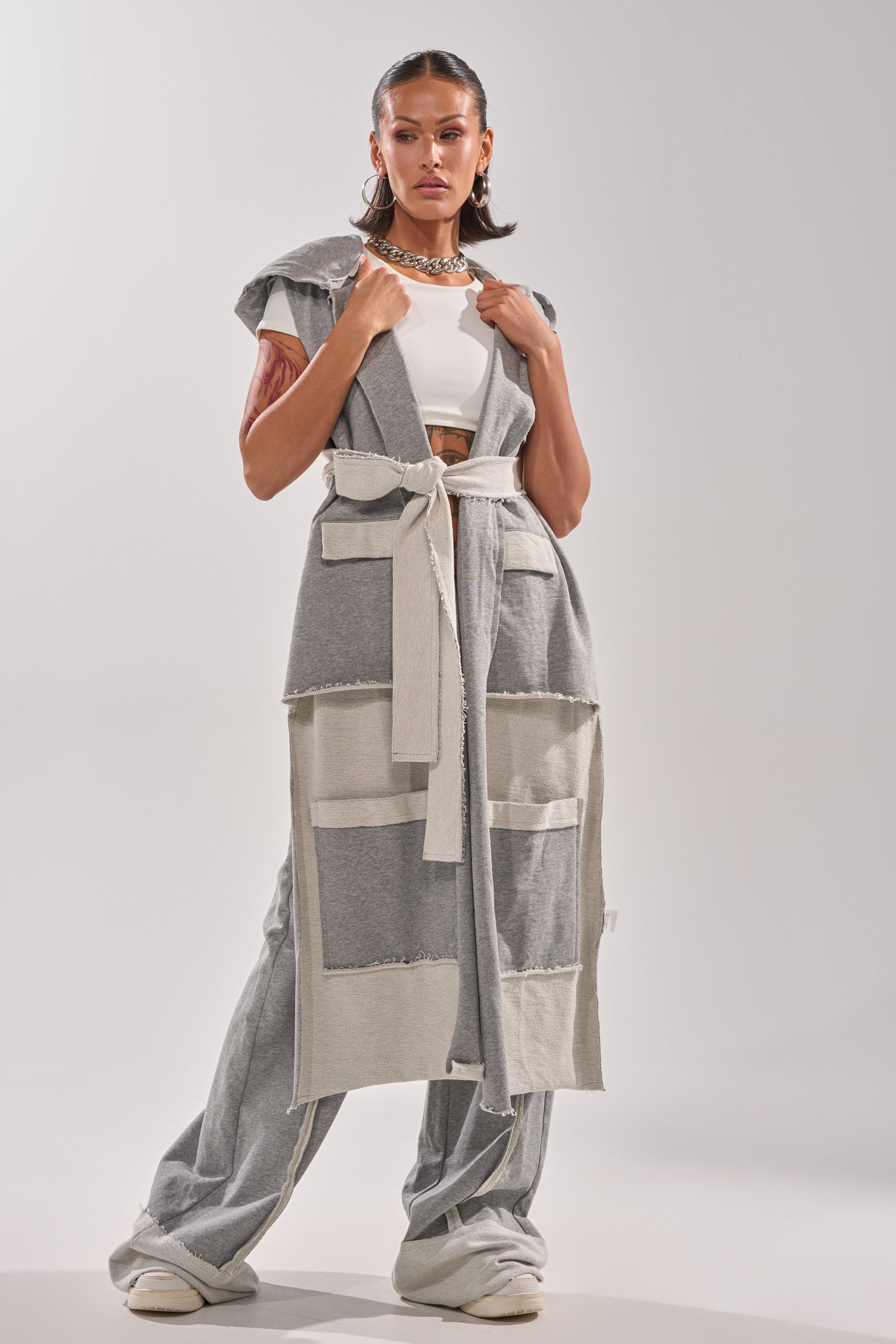 A woman exudes confidence in the IM UP NEXT DUSTER, paired with a white crop top, wide-legged gray pants, chunky white sneakers, hoop earrings, and a chain necklace for a striking statement ensemble.