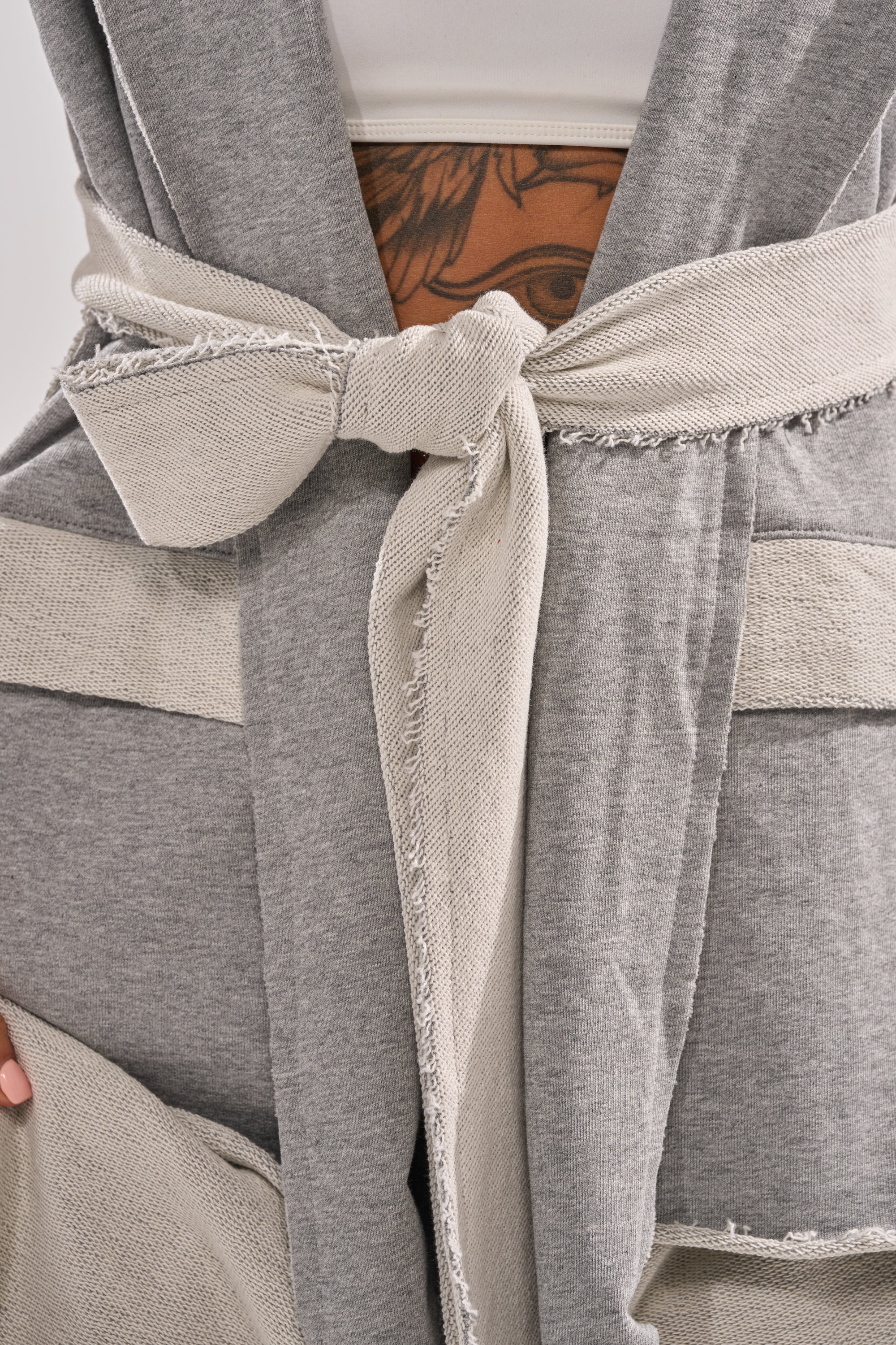 Close-up of a person wearing the IM UP NEXT DUSTER, a gray and beige two-tone piece with a fabric belt at the waist. A tattoo is visible above the belt as one hand holds the duster's hem.