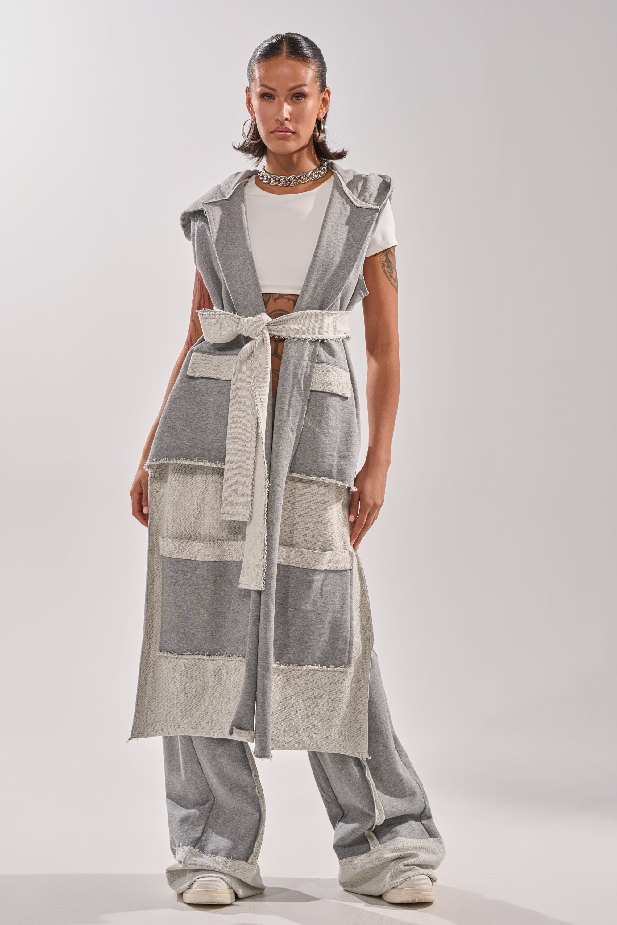A woman faces forward, wearing the IM UP NEXT DUSTER—a long, sleeveless gray vest with large pockets and a belt—styled with wide-leg gray pants, a white crop top, chunky necklace, and white sneakers against a plain light background.