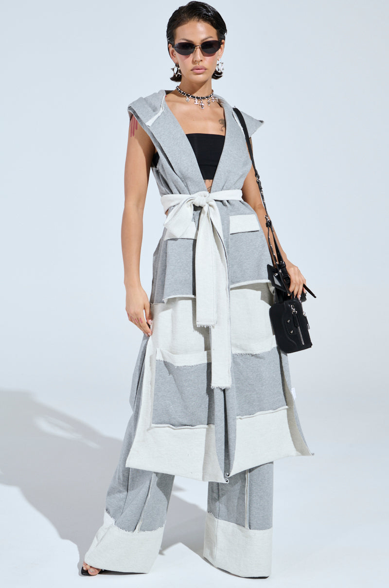 A person confidently poses in a fashionable two-tone patchwork outfit, featuring our IM UP NEXT DUSTER—a sleeveless hoodie with a plunging neckline—paired with wide-leg pants and sunglasses, accessorized with a black handbag and chunky necklace.