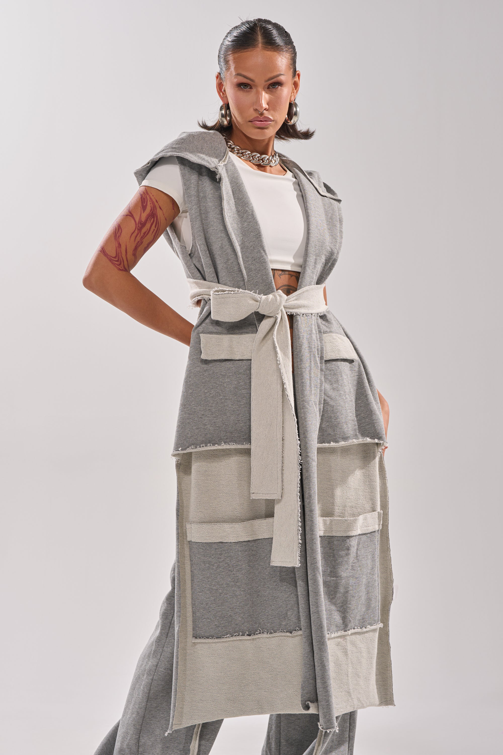 A woman poses confidently in the IM UP NEXT DUSTER from AKIRA Label, a sleeveless two-tone design, styled over a white crop top and gray pants. She sports slicked-back hair, an arm tattoo, and chunky jewelry.