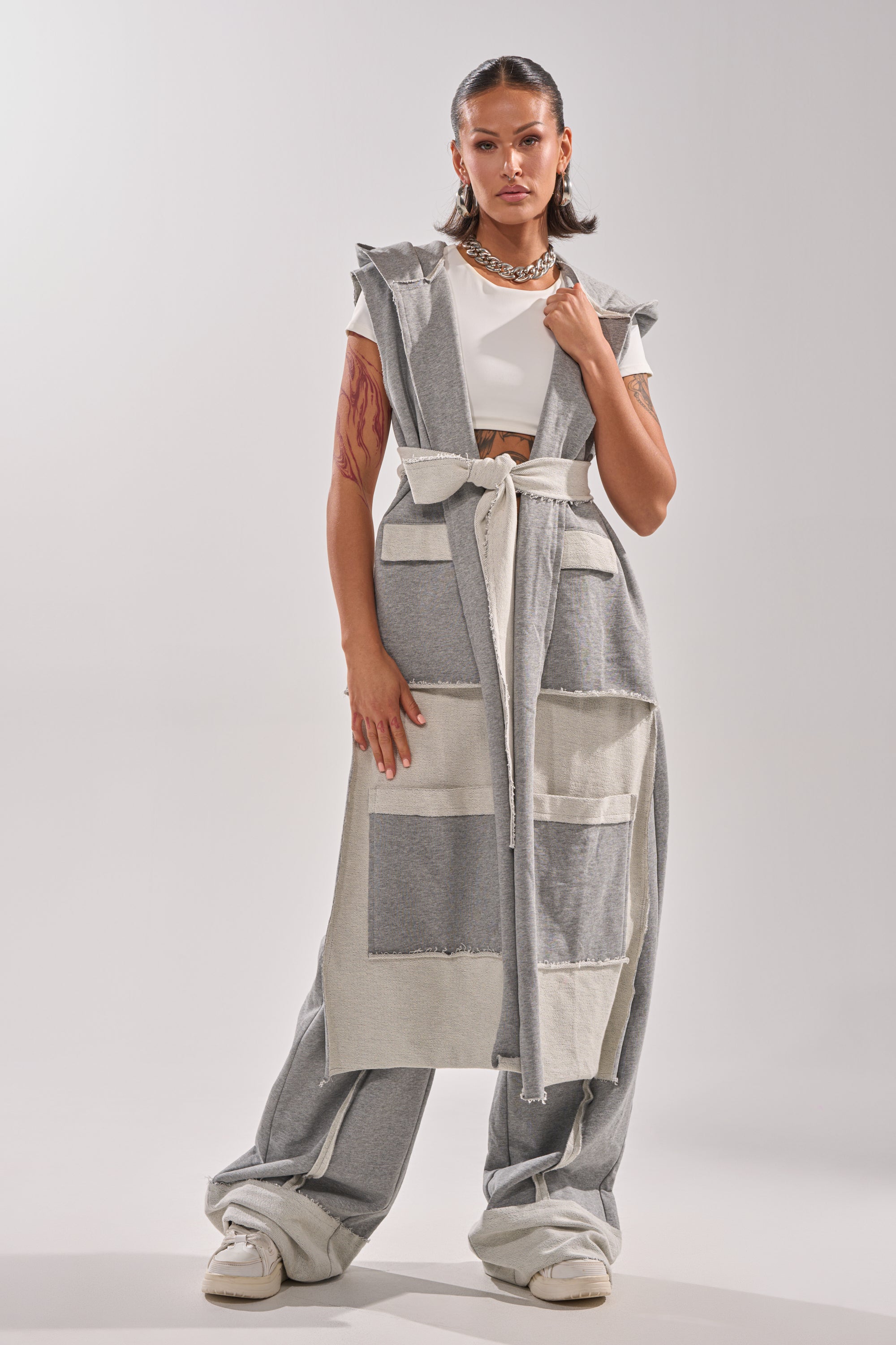 A woman stands confidently in the IM UP NEXT DUSTER, a grey two-tone sleeveless hooded vest with patch pockets, layered over matching wide-leg pants. She pairs it with a white crop top, sneakers, and a chunky chain necklace, hair slicked back.