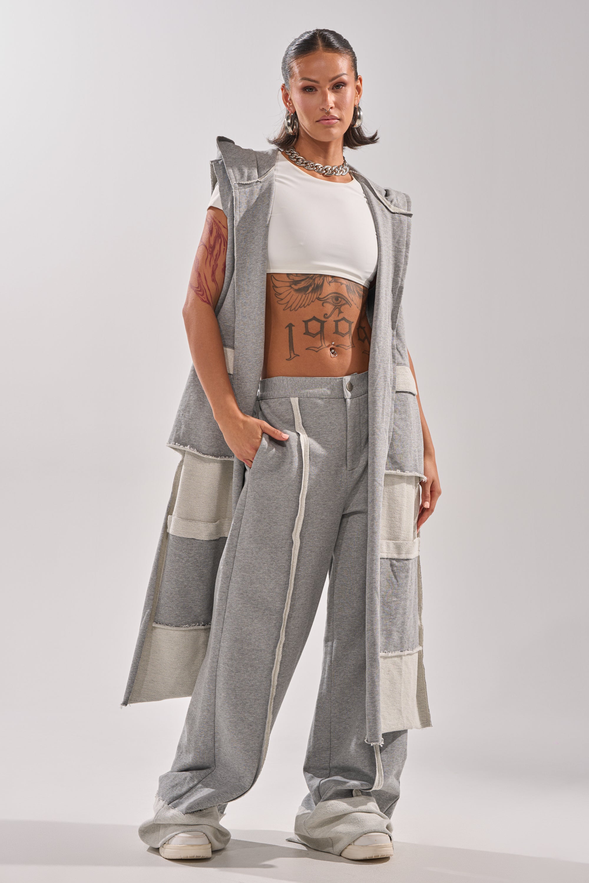 A woman confidently wears the IM UP NEXT DUSTER, a long, sleeveless gray duster paired with matching wide-leg pants, a white crop top, white slippers, a silver necklace, and displays tattoos on her torso and arm.