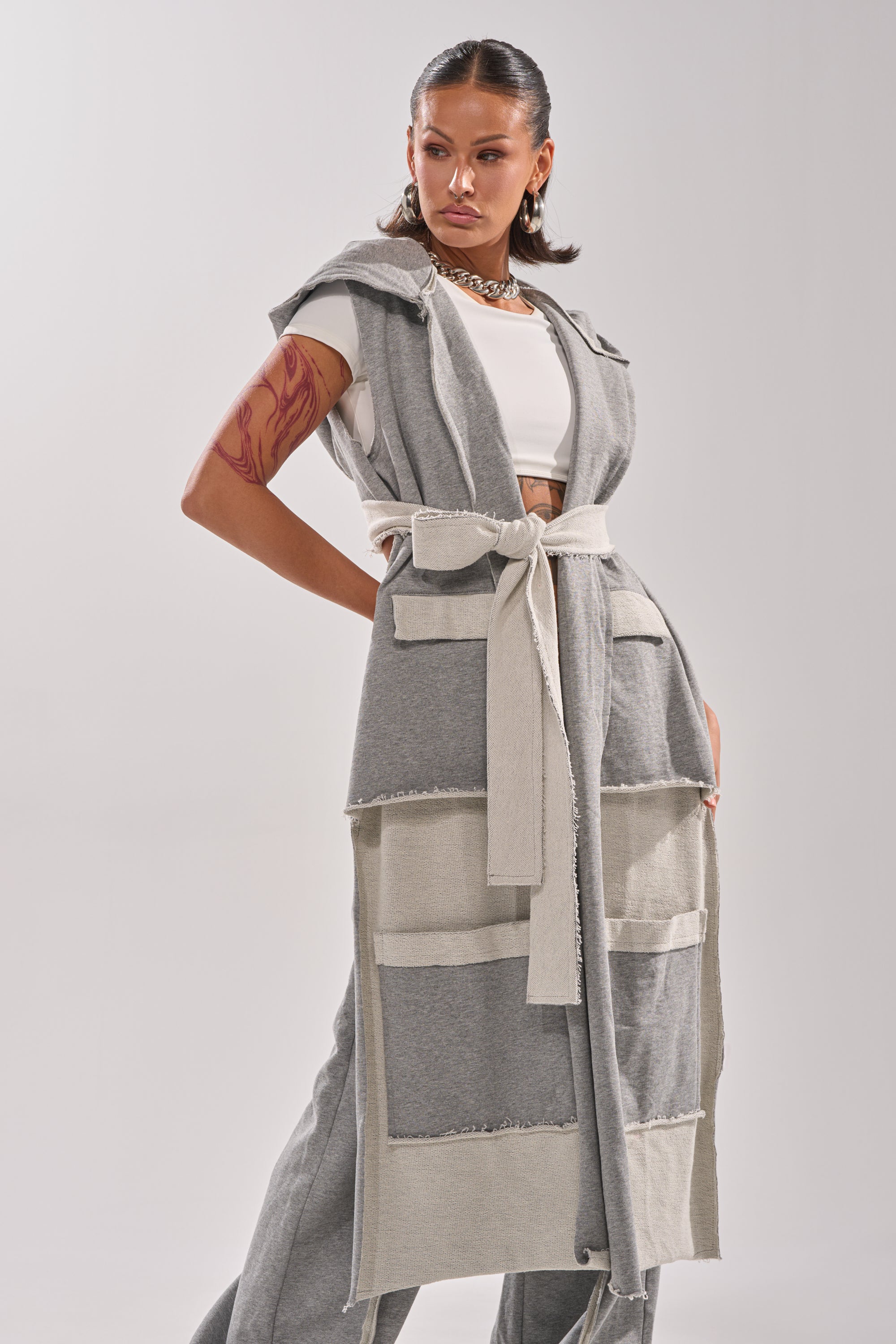 A woman exudes confidence in the IM UP NEXT DUSTER—a gray, sleeveless piece with large pockets and a belted waist, styled over a white crop top and pants. A bold arm tattoo and chunky chain necklace complete her standout look.
