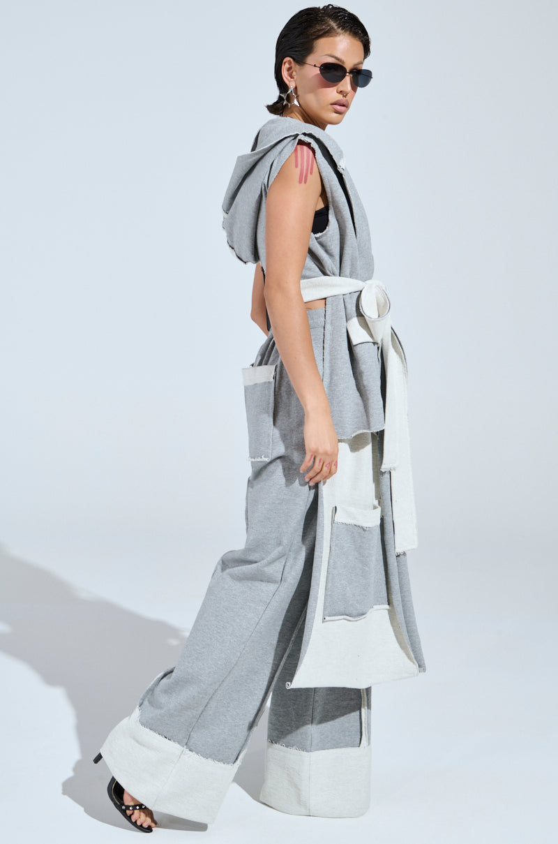 A person poses in a gray sleeveless IM UP NEXT DUSTER with a plunging neckline and large pockets, paired with wide-legged pants featuring contrasting cuffs and black high-heeled sandals, against a light background.