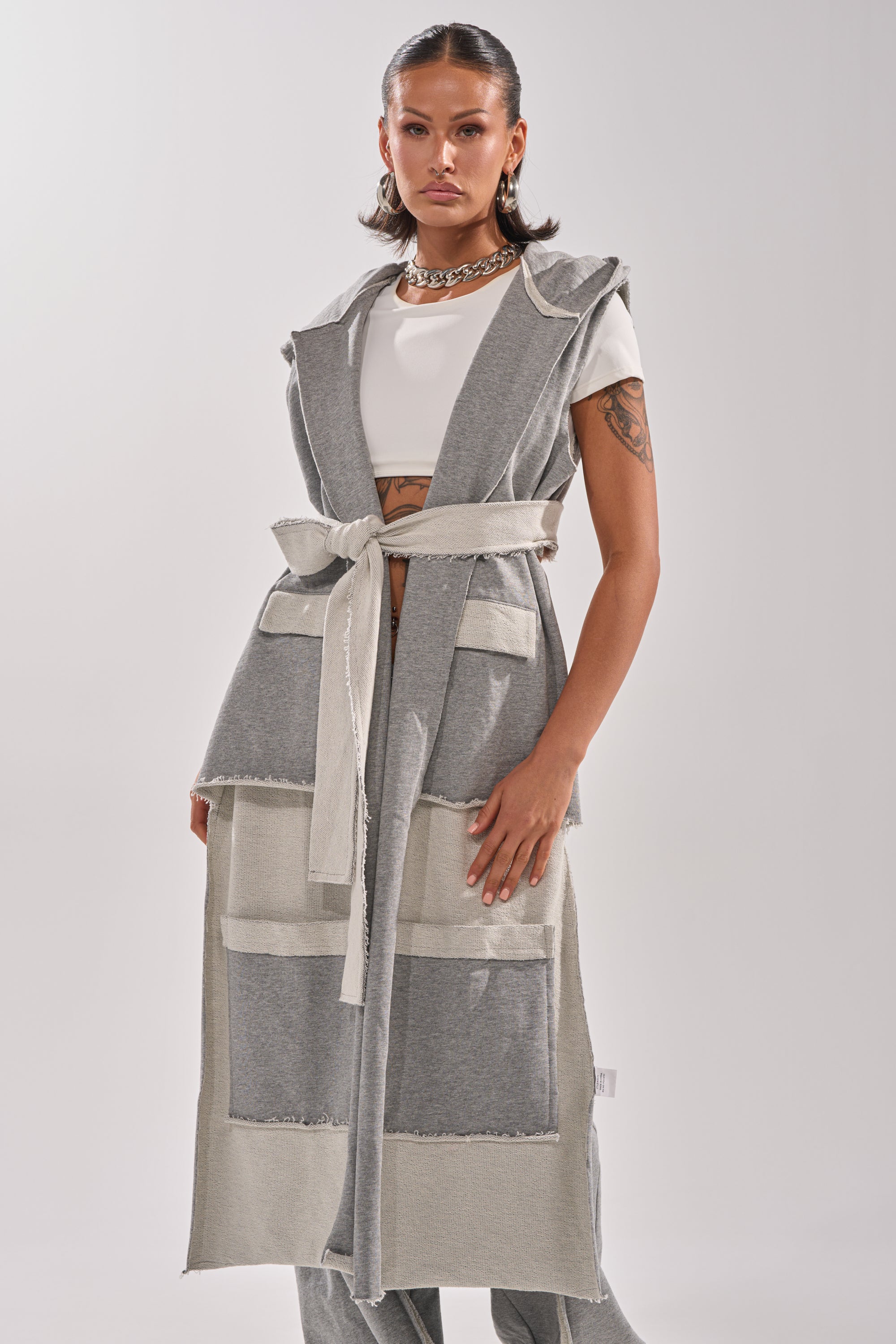 A woman with dark, wavy hair styles the IM UP NEXT DUSTER—a long, sleeveless piece with large front pockets and a matching belt—over a white crop top, finishing the look with hoop earrings and a chunky chain necklace.