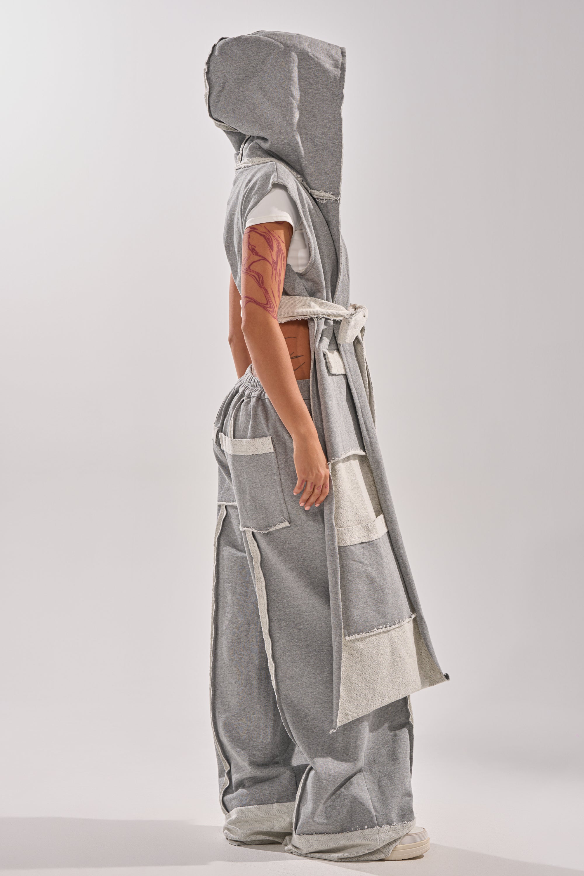 A person stands in profile wearing oversized patchwork gray and white pants paired with the IM UP NEXT DUSTER, showcasing a modern, avant-garde style with a visible arm tattoo.