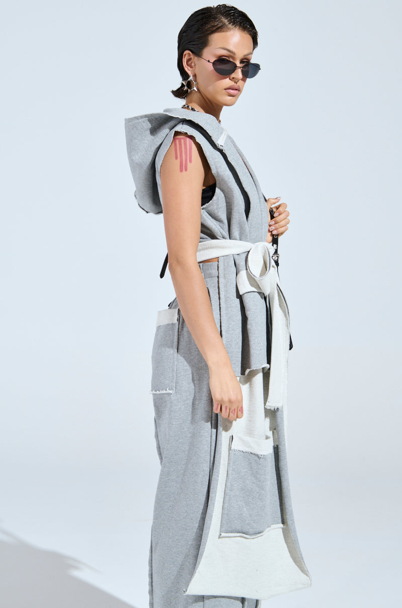 A person confidently wears the stylish "IM UP NEXT DUSTER," a sleeveless gray outfit with a two-tone design, large pockets, and wrap-around belt. The plain, light-shaded background enhances the ensemble’s chic modern flair.