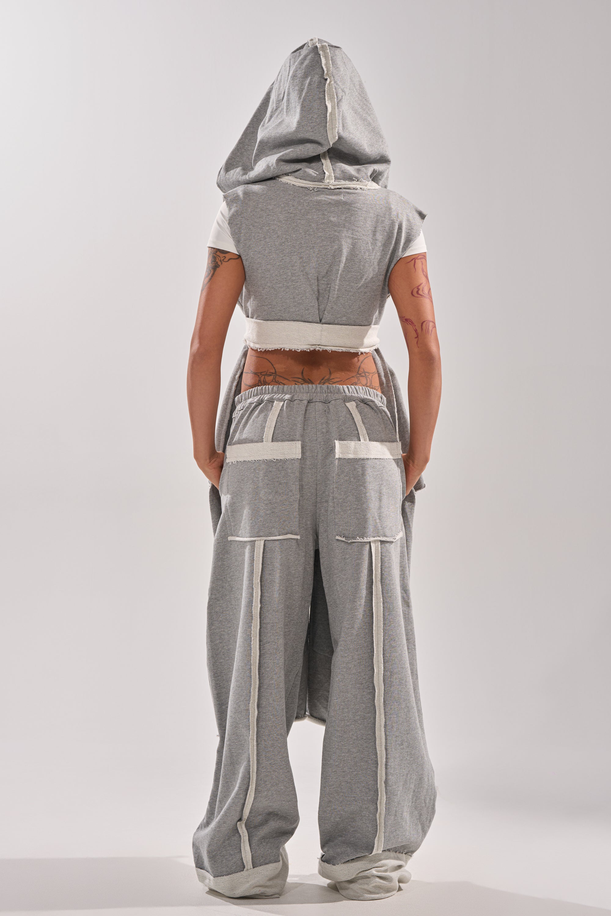 A person with tattooed arms faces away, wearing the IM UP NEXT DUSTER— a gray sleeveless hooded top with exposed seams— and matching wide-leg pants with white trim and large back pockets, set against a plain light background.