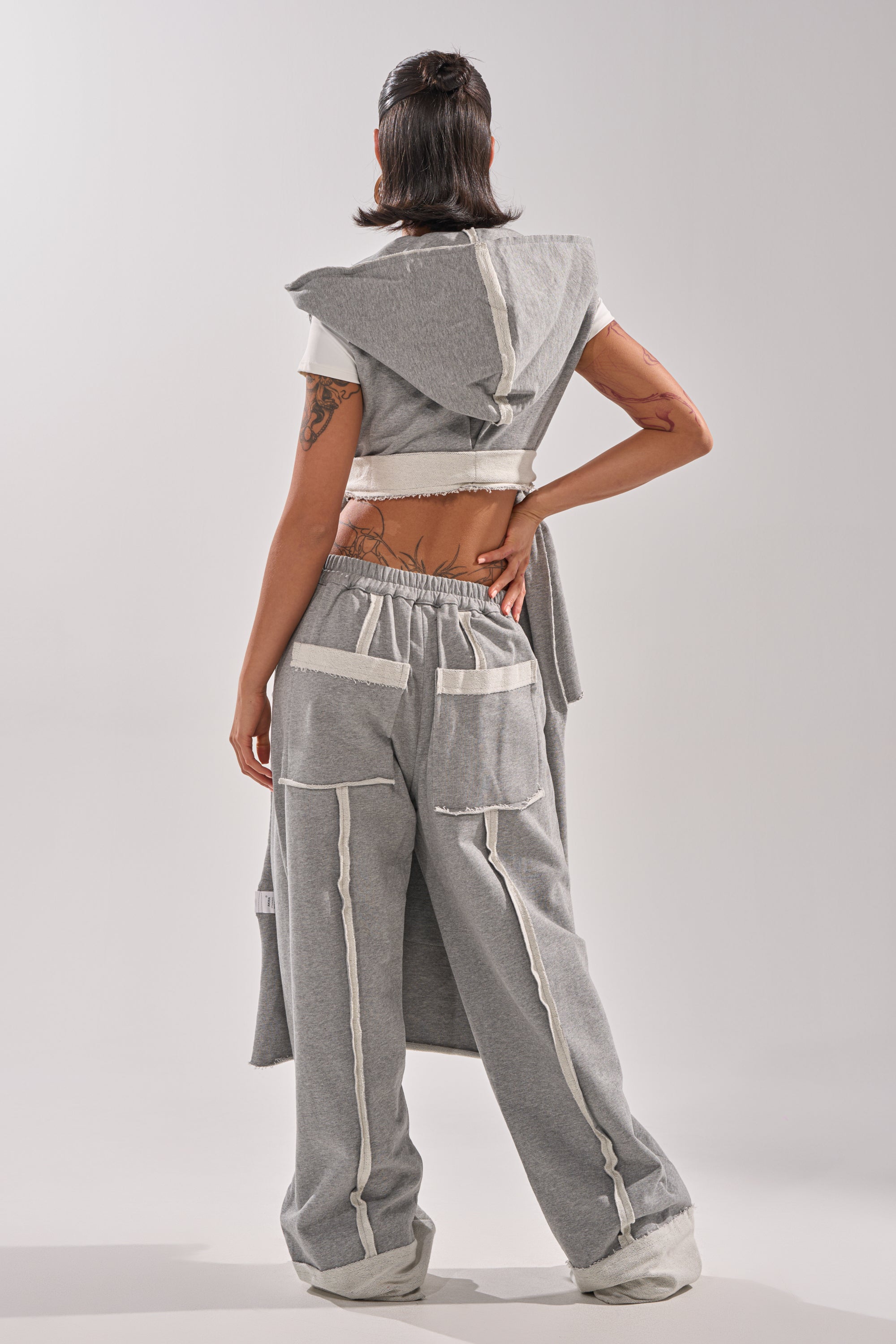 A person stands facing away, wearing the IM UP NEXT DUSTER layered over a matching gray outfit of wide-leg pants and a short-sleeve top with exposed seams, white trim, and large back pockets for a modern, effortless vibe.