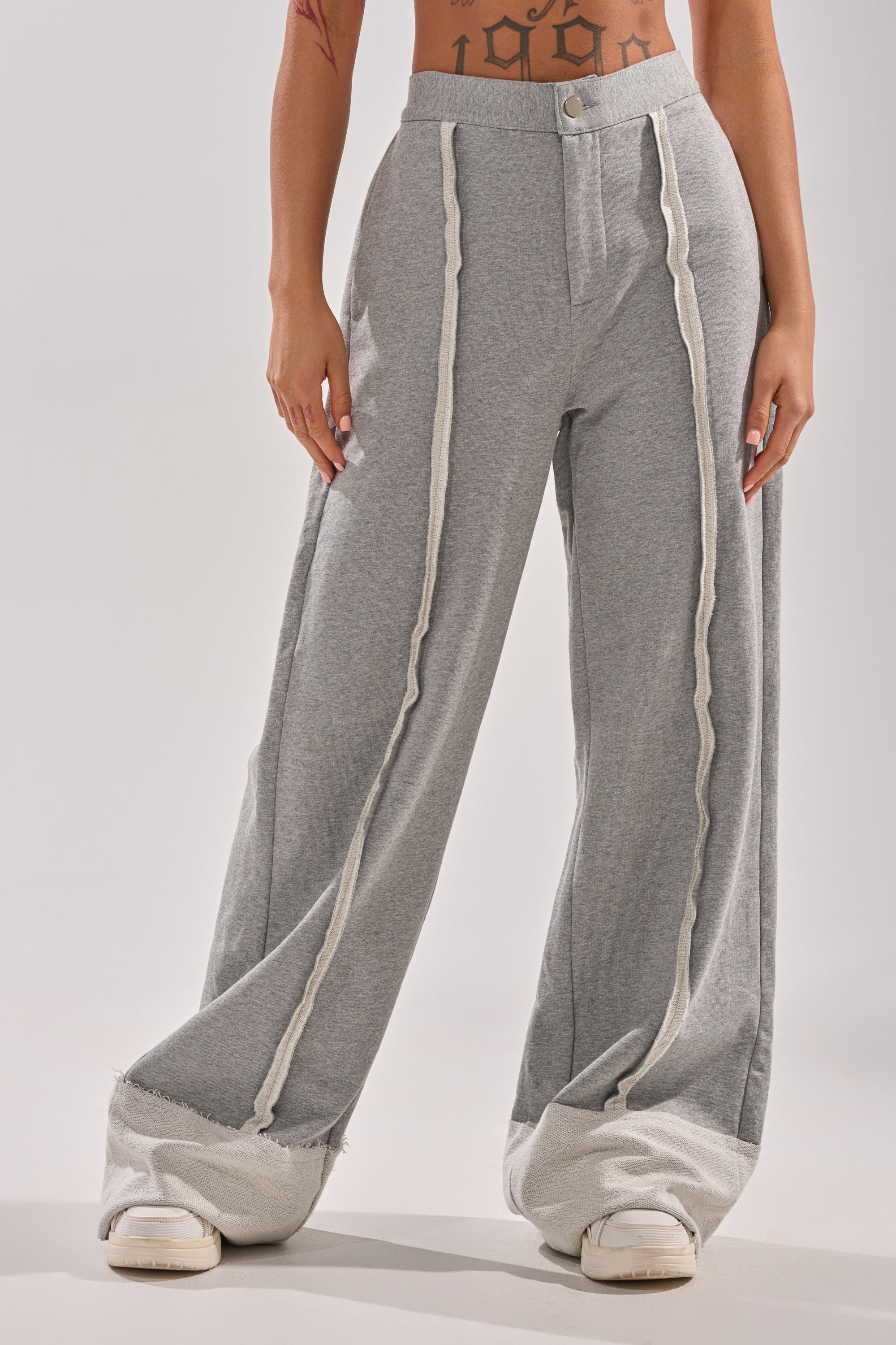 A person wears AKIRA Label IM UP NEXT TROUSER—loose-fitting, high-rise gray pants with white textured trim front and hem—paired with white sneakers, revealing a partially visible midriff and tattooed arms.