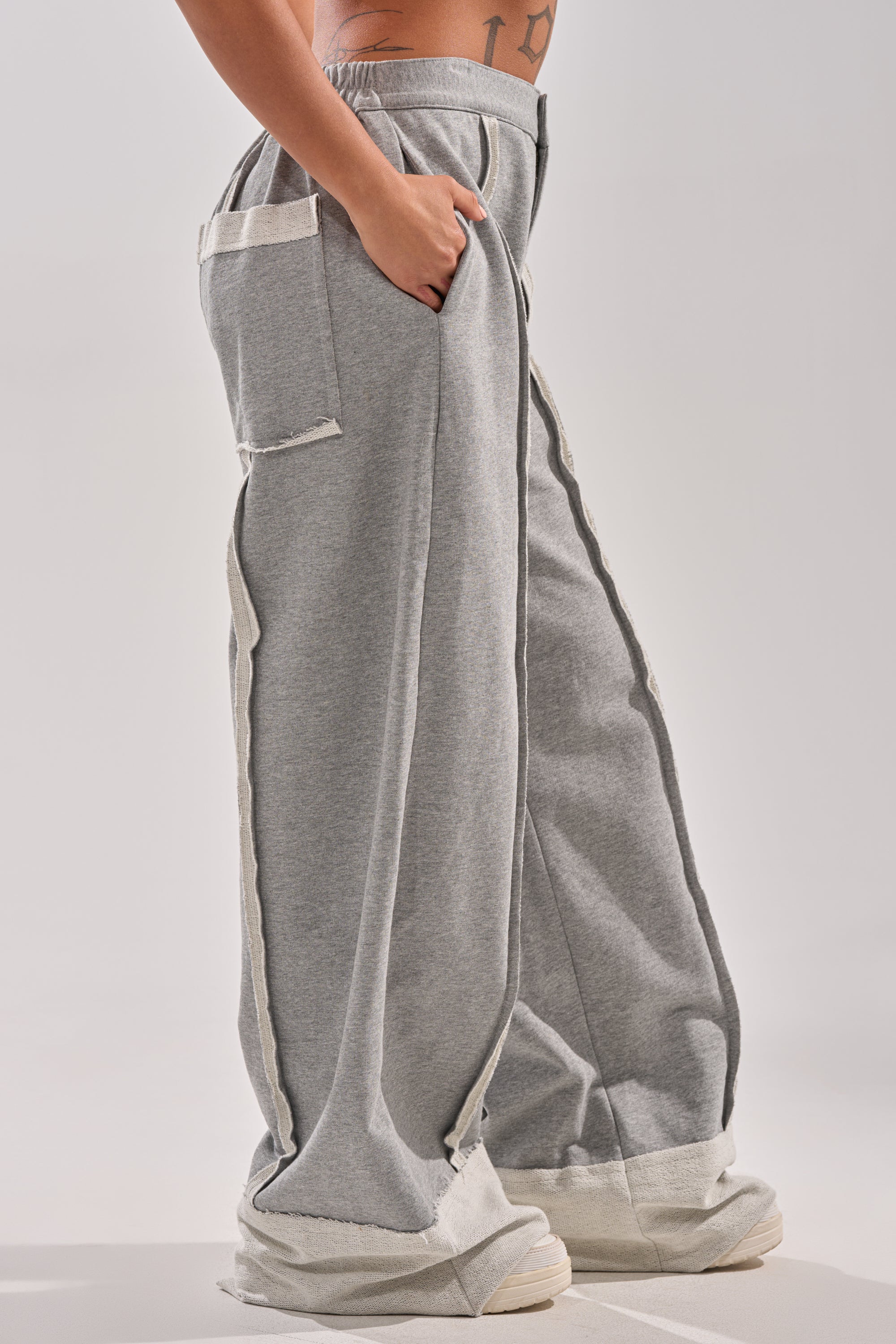 A person stands sideways against a plain light background, with only the lower torso and legs visible, wearing IM UP NEXT TROUSER: loose-fitting gray cotton trousers featuring large pockets and visible seams. One hand is in a pocket.