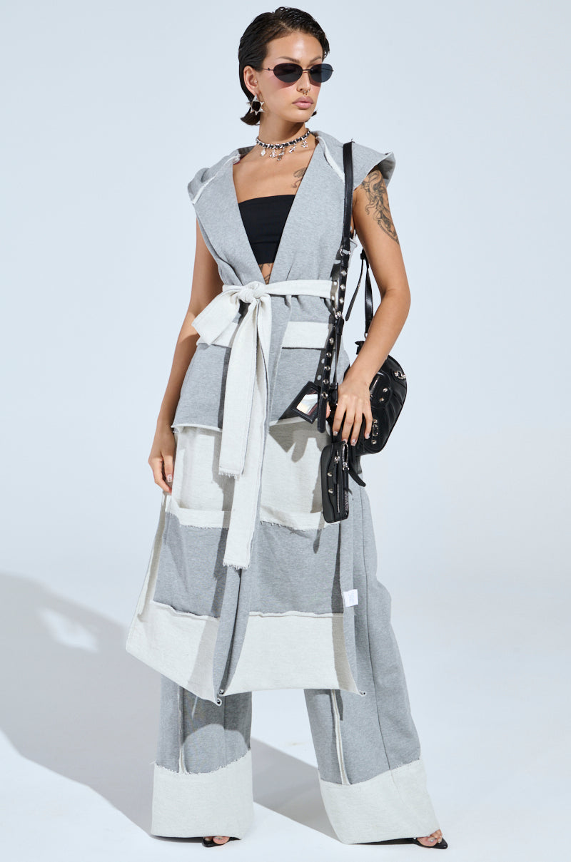 A person poses confidently in a stylish outfit, featuring sunglasses, a sleeveless gray layered vest with white trim, and a black top with shirred elastic panels. They pair these with the IM UP NEXT TROUSER and hold a black handbag. Their look is completed by short hair and a necklace against a plain background.