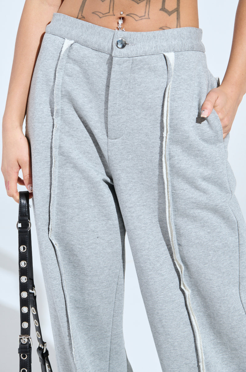 A person wears the IM UP NEXT TROUSER in grey, featuring raw hems and a button with a high-rise fit. They're holding a black strap with metal rings, revealing part of their midriff and showing a navel piercing and tattoo.