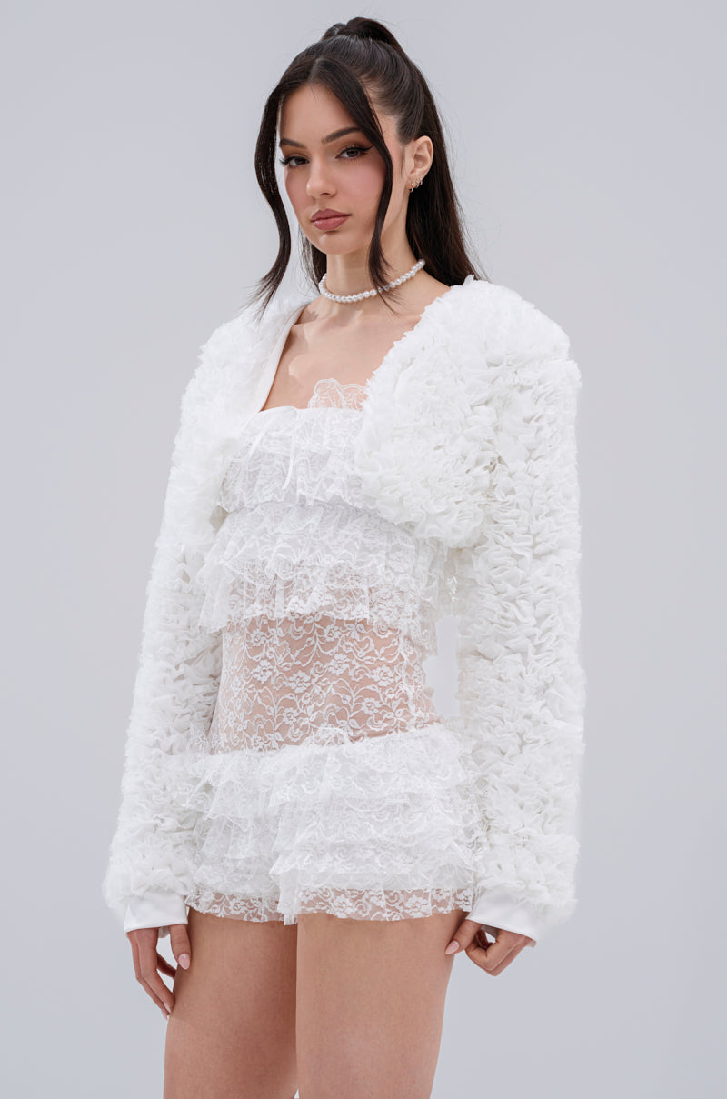 A woman with long dark hair in a half-up style and a pearl necklace stands against a grey background, wearing the PLUME RUFFLE BOLERO over a sheer white lace dress. She looks directly at the camera.