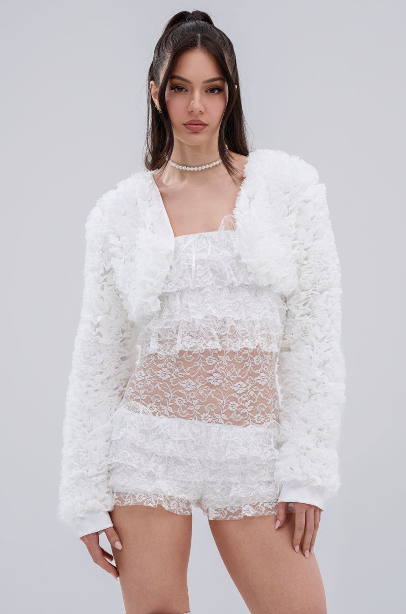 A woman with long dark hair in loose waves wears the PLUME RUFFLE BOLERO over a sheer white lace dress, paired with a pearl necklace, standing against a plain light gray background.