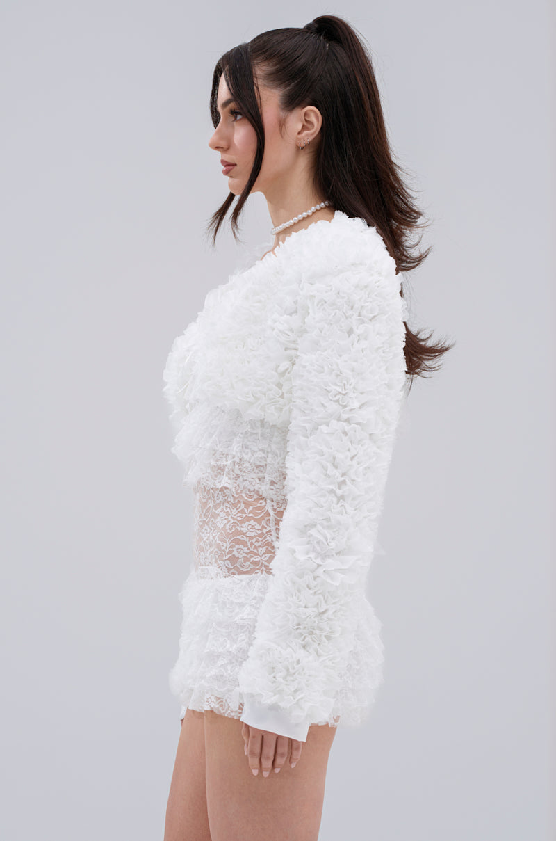 A woman in profile stands against a plain background, wearing the white PLUME RUFFLE BOLERO with voluminous ruffles over a lace dress, her dark hair styled in a high ponytail and accessorized with a pearl necklace.