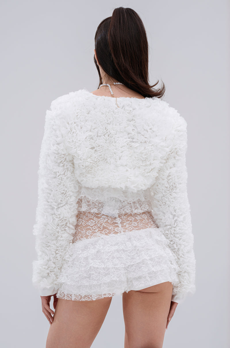 A person with long dark hair in a high ponytail wears the PLUME RUFFLE BOLERO, a white cropped jacket with voluminous ruffles, and a lace skirt, seen from the back against a plain light gray background.