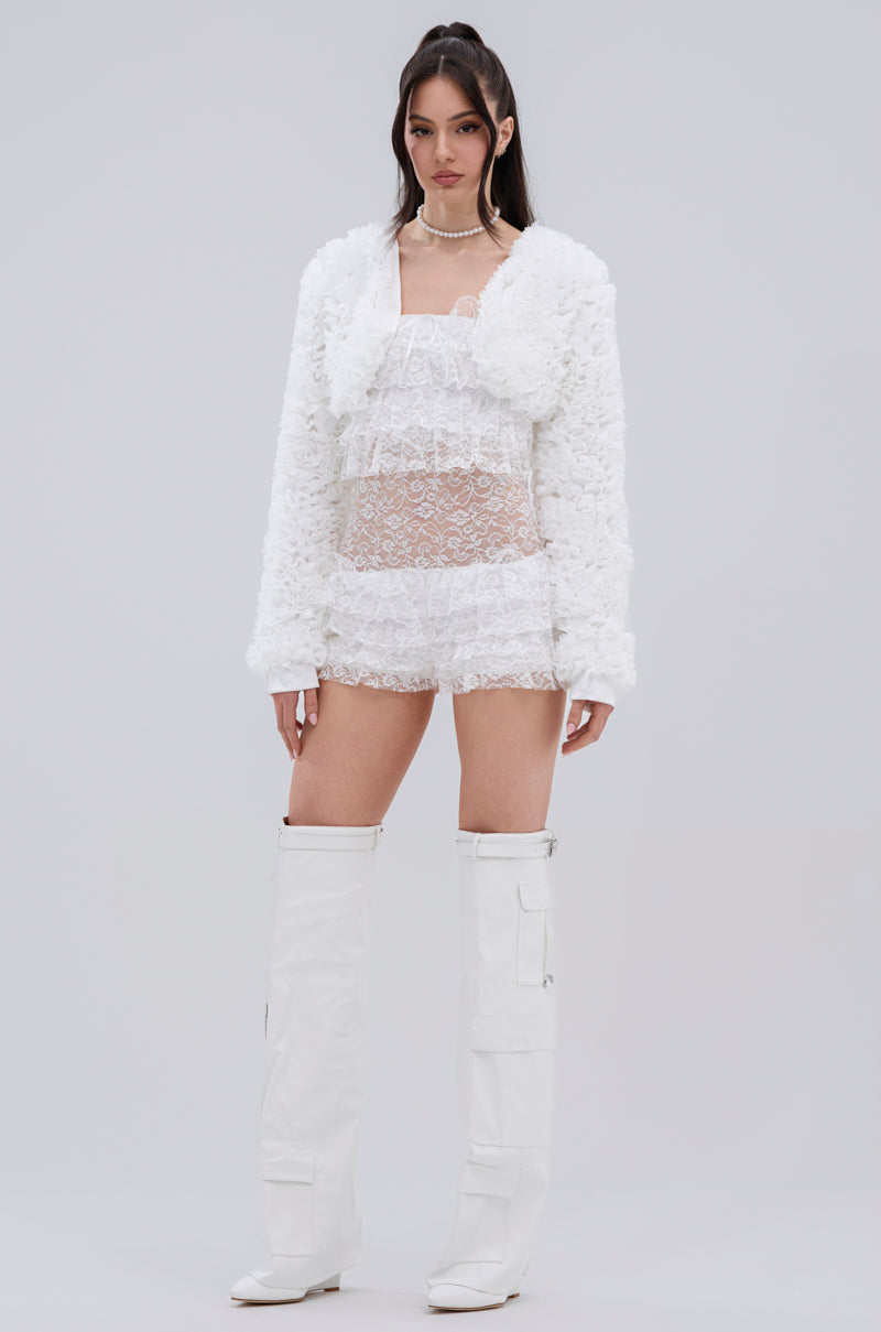A woman poses against a plain background in the PLUME RUFFLE BOLERO, layered over a sheer lace dress with tall boots. Her long dark hair and neutral expression enhance the bolero’s subtle elegance.