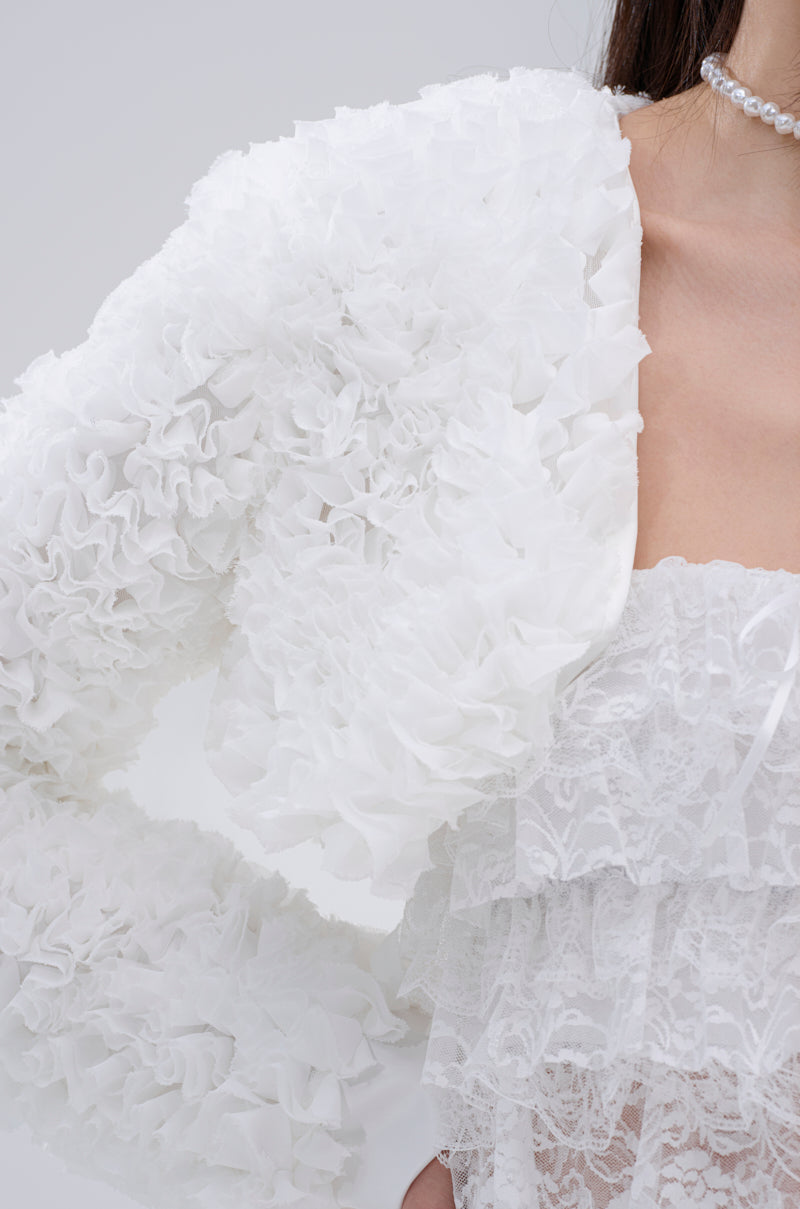 A close-up shows a person wearing the PLUME RUFFLE BOLERO, a white voluminous ruffle jacket over a lace top with a pearl necklace. Only part of the face and upper body are visible against a light gray background.