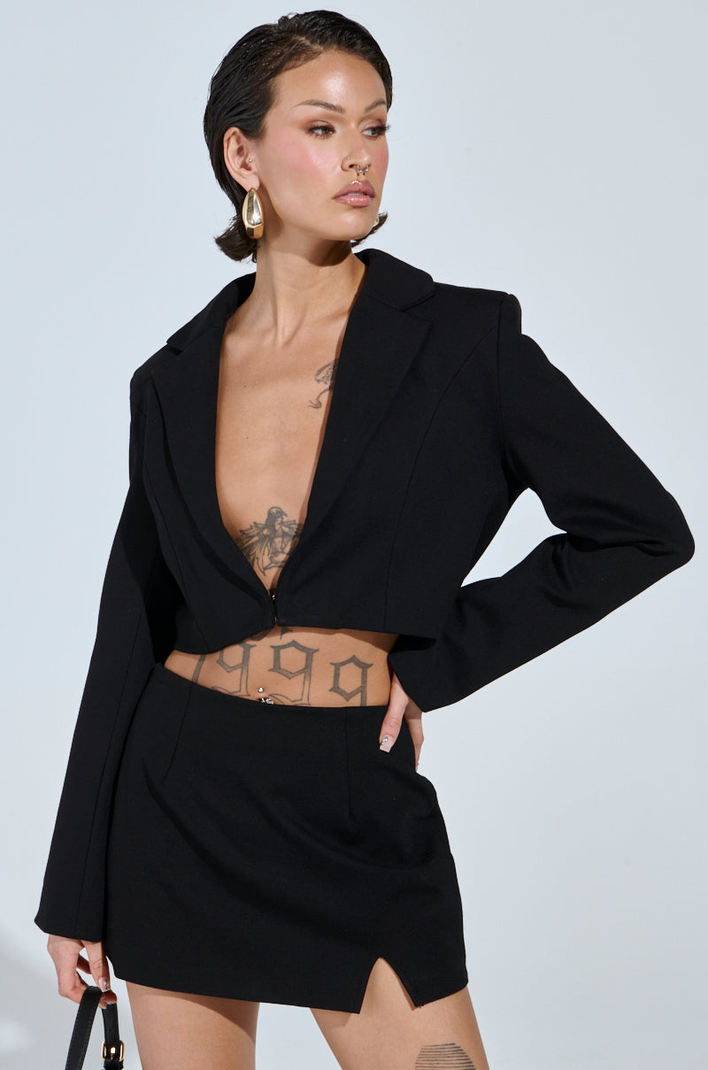 Wearing an AKIRA Label NEW CLASSIC CROPPED BLAZER, a person with short dark hair and prominent torso tattoos poses sideways in a black tailored, deep V-neck style. They pair it with a matching high-waisted mini skirt and hold a small handbag neutrally—a true wardrobe staple.