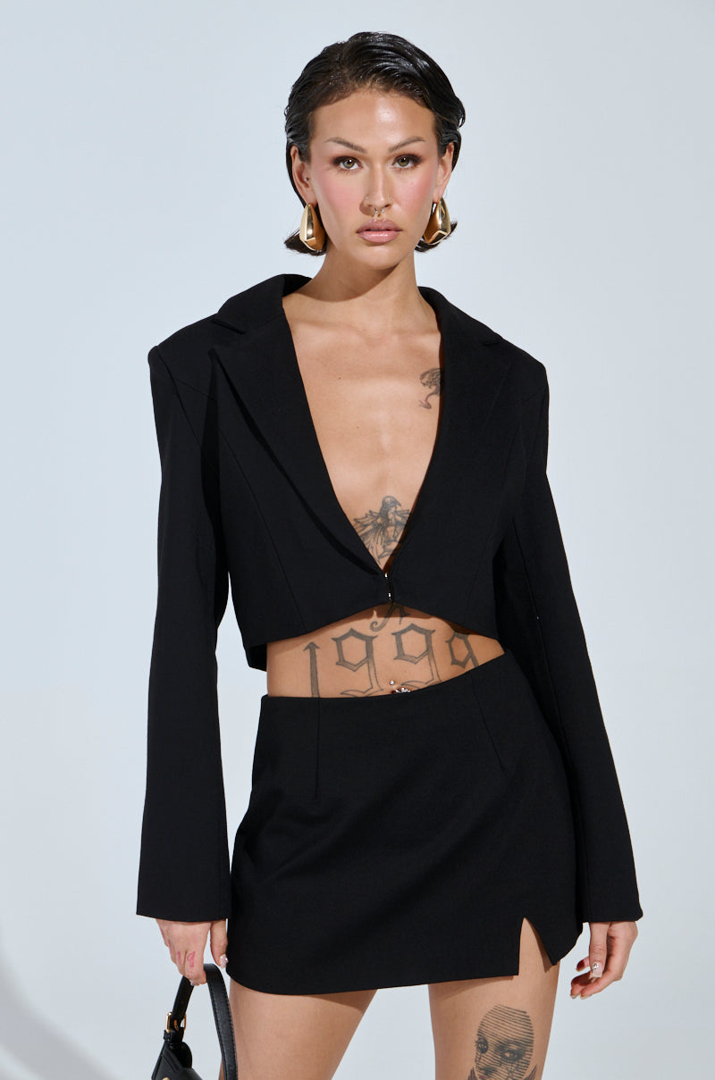 A person with short dark hair wears the AKIRA Label NEW CLASSIC CROPPED BLAZER paired with a matching skirt. Tattoos adorn their chest and upper legs as they hold a bag, complemented by large gold hoop earrings, against a plain background.