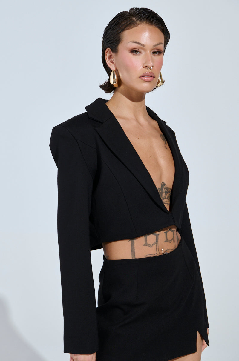 A person with short, slicked-back hair wears a NEW CLASSIC CROPPED BLAZER—a wardrobe staple—with an AKIRA Label skirt. Visible tattoos, hoop earrings, and subtle makeup enhance their modern, chic style against a plain background.