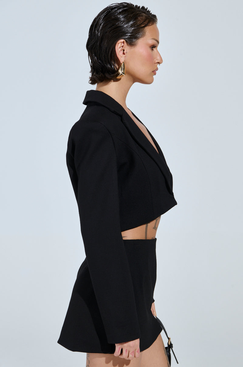 A person with short, slicked-back hair wears the AKIRA Label's NEW CLASSIC CROPPED BLAZER and a black skirt. They stand in profile against a plain background, accessorized with large hoop earrings—a true wardrobe staple.