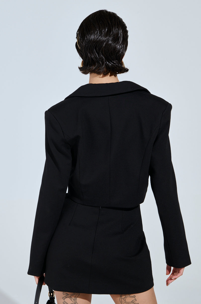 A person with short, dark hair stands facing away, highlighting the NEW CLASSIC CROPPED BLAZER and matching skirt—essentials from the AKIRA Label. Tattoos decorate their legs as they hold a handbag against a plain, light background.