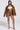 A confident woman wears a BIRDIE OVERSIZED BOMBER, its quilted interior visible beneath brown leather. She pairs it with a white tube top, matching shorts, and mixed-pattern high heels against a plain background.