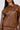 A person wears the BIRDIE OVERSIZED BOMBER, a brown faux leather jacket with a zipper, large chest pockets, and a quilted interior. It's paired with matching pants. The person has long dark hair and hoop earrings, showcasing their stylish look from the upper torso up.