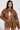 A person with long dark hair wears a brown BIRDIE OVERSIZED BOMBER and matching shorts over a white top, posing confidently with one hand on their waist. The quilted interior adds texture against a neutral background.
