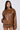 Confidently standing, the woman dons a BIRDIE OVERSIZED BOMBER in brown leather with a quilted interior and matching shorts. Her long dark hair flows down, complemented by hoop earrings. The light gray background highlights her chic style.