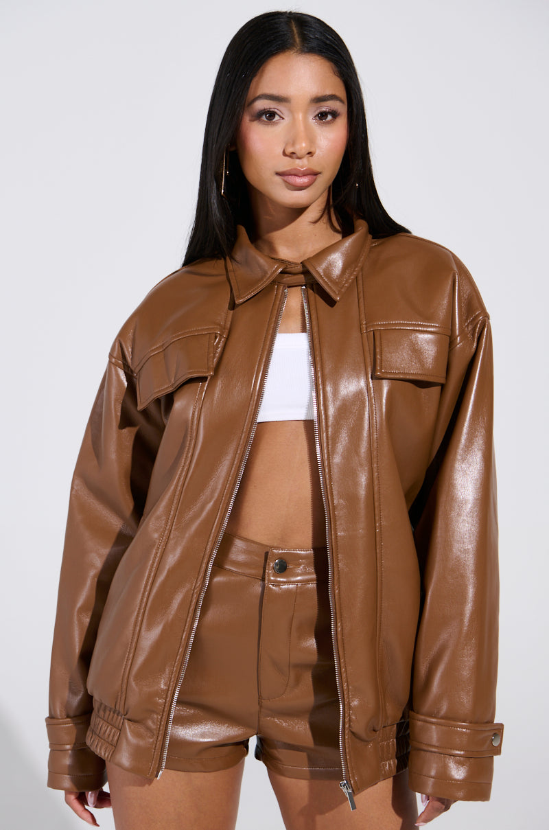 A person with long hair is wearing the BIRDIE OVERSIZED BOMBER, featuring a chic faux leather exterior and quilted interior. It's paired perfectly with matching shorts, hoop earrings, and a white top. The plain, light-colored backdrop accentuates their bold fashion statement.