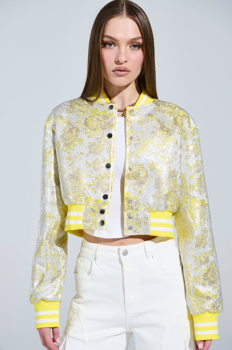 A person with long hair is wearing a SOLEIL RHINESTONE BROCADE BOMBER in yellow and silver sequins over a white top and pants. The jacket features button closures and striped cuffs, with the plain light background enhancing the chic look.
