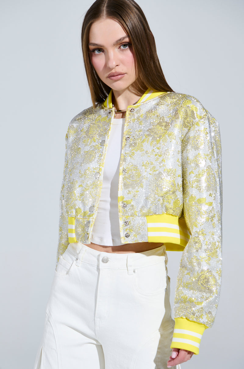 A person with long brown hair, dressed in a SOLEIL RHINESTONE BROCADE BOMBER featuring a shiny silver and yellow floral pattern with rhinestone highlights, wears a white crop top and pants in front of a plain light-colored background. The jacket has yellow-striped cuffs and hem.