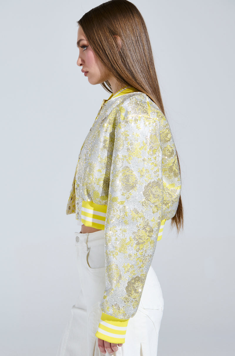 A person with long brown hair stands in profile, wearing the SOLEIL RHINESTONE BROCADE BOMBER in sparkling silver and yellow, featuring rhinestone brocade. The yellow and white striped cuffs complement high-waisted white pants against a plain light gray background.