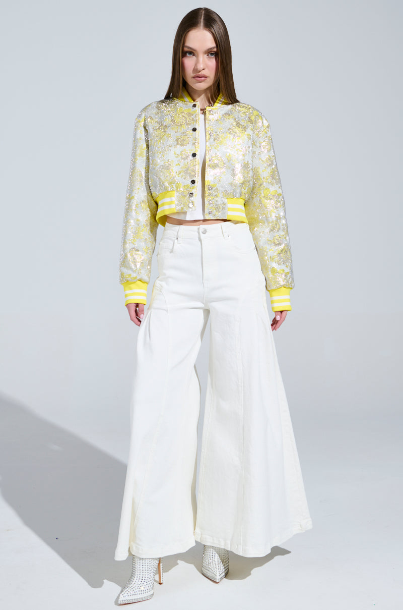 Against a plain background, a person exudes confidence in the SOLEIL RHINESTONE BROCADE BOMBER, featuring floral patterns with silver snap-buttons. They pair it seamlessly with wide-leg white pants and textured ankle boots, complemented by their long hair for a stylish ensemble.