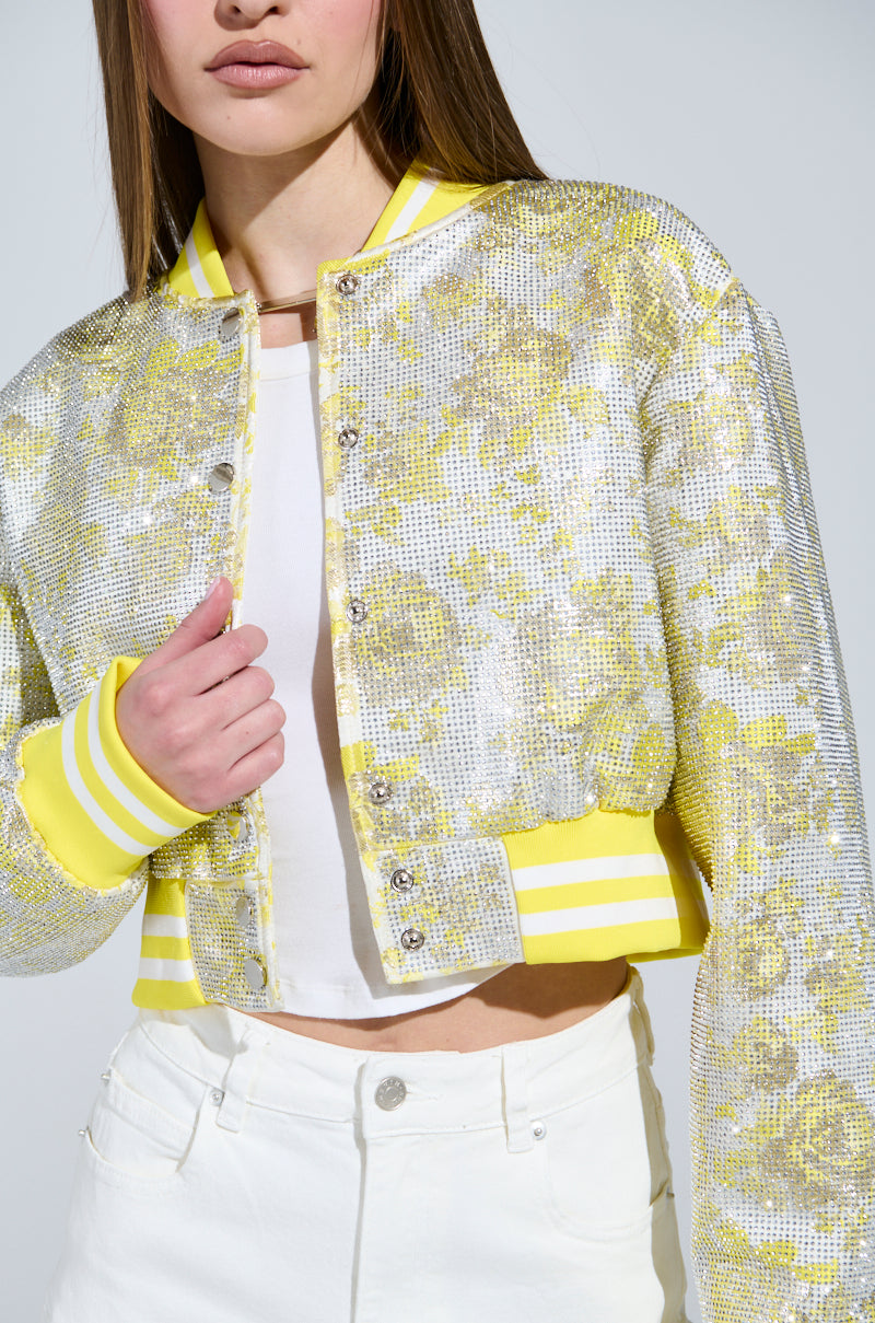 The individual wears the SOLEIL RHINESTONE BROCADE BOMBER, a shiny silver and yellow floral-patterned jacket featuring rhinestone brocade with silver snap-buttons at the cuffs and collar. Beneath, a white top and white pants complement the look against a plain backdrop.