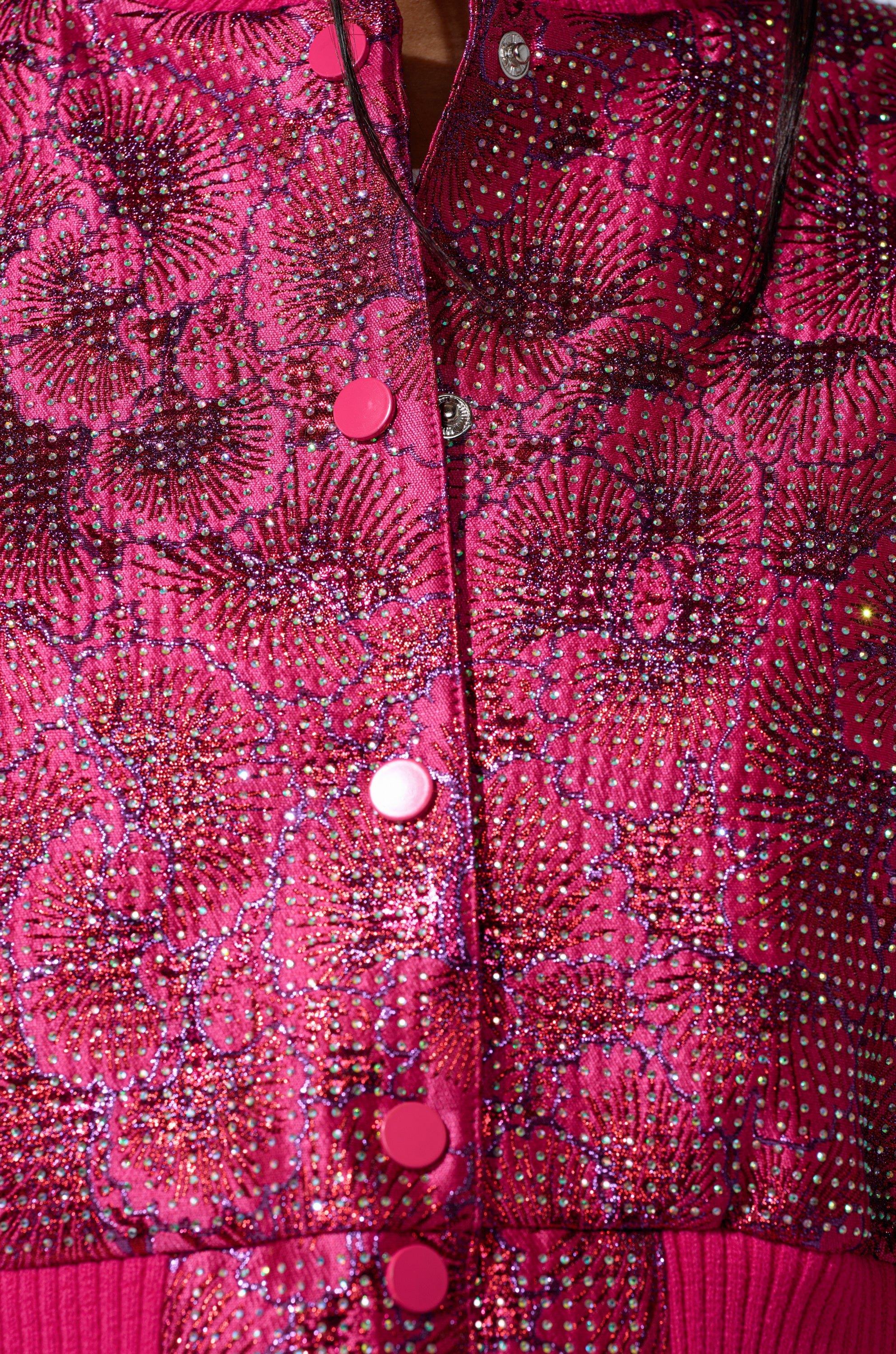 A close-up of the FLEUR RHINESTONE BROCADE BOMBER, featuring shiny, textured floral brocade with sparkling rhinestone embellishments, pink buttons down the center, and intricate floral patterns.