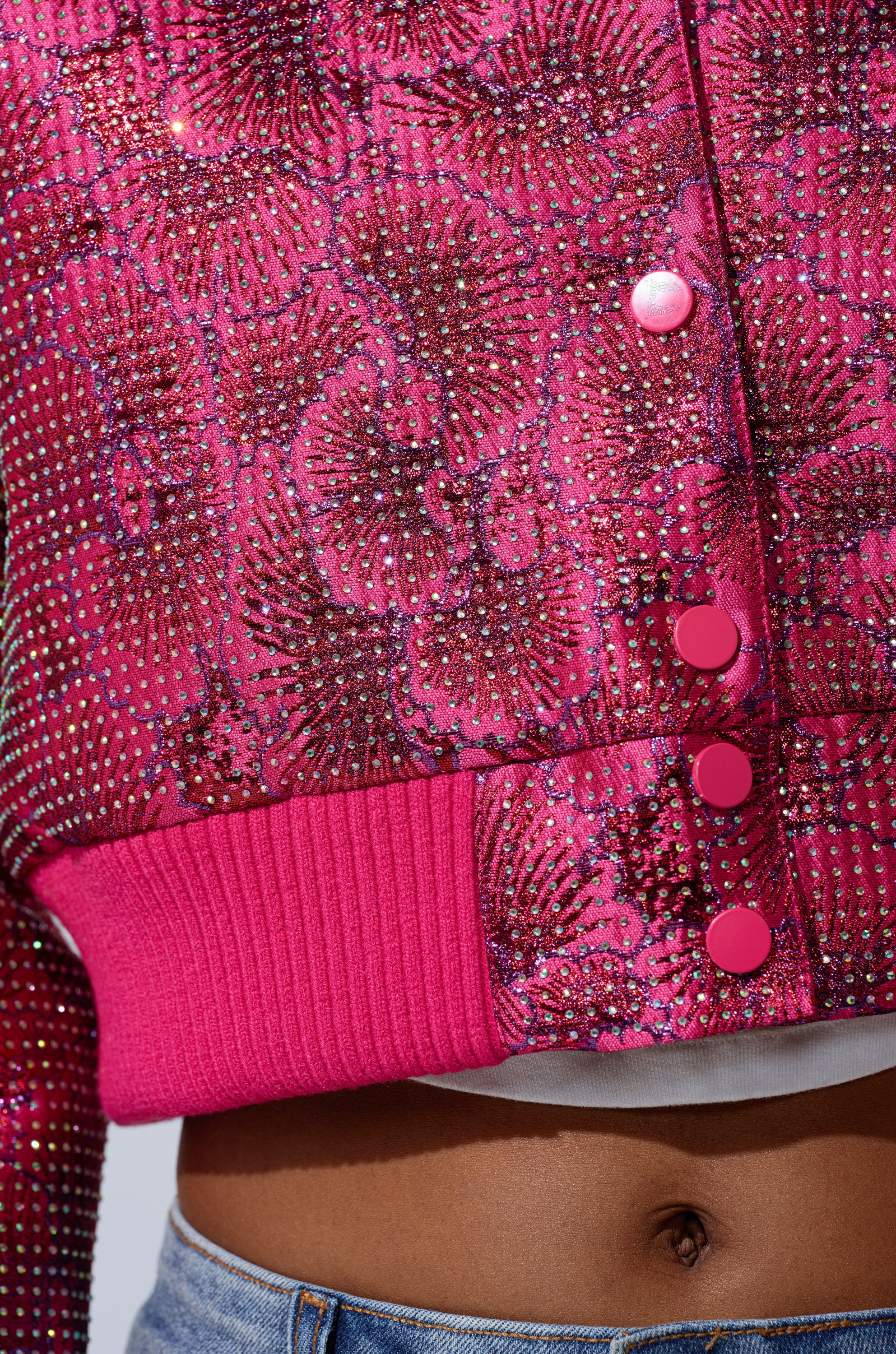 Close-up of the FLEUR RHINESTONE BROCADE BOMBER in pink, showcasing an intricate rhinestone pattern, ribbed hem, and pink buttons. Layered over a white top with blue jeans for a chic, slightly midriff-baring look.